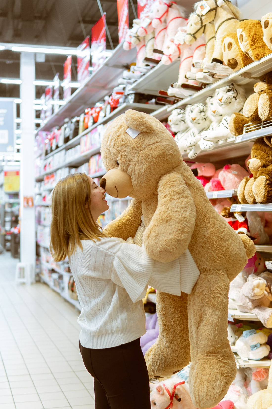 Woman browsing plush toys on store shelves while holding a large teddy bear