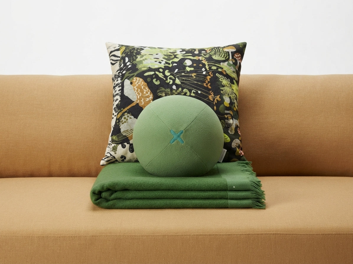 Back of the green olive plush pillow with cross-stitched seam detail, on a tan sofa with a green throw