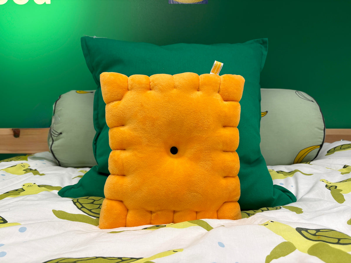Cheese cracker plush pillow propped against a green cushion on a bed